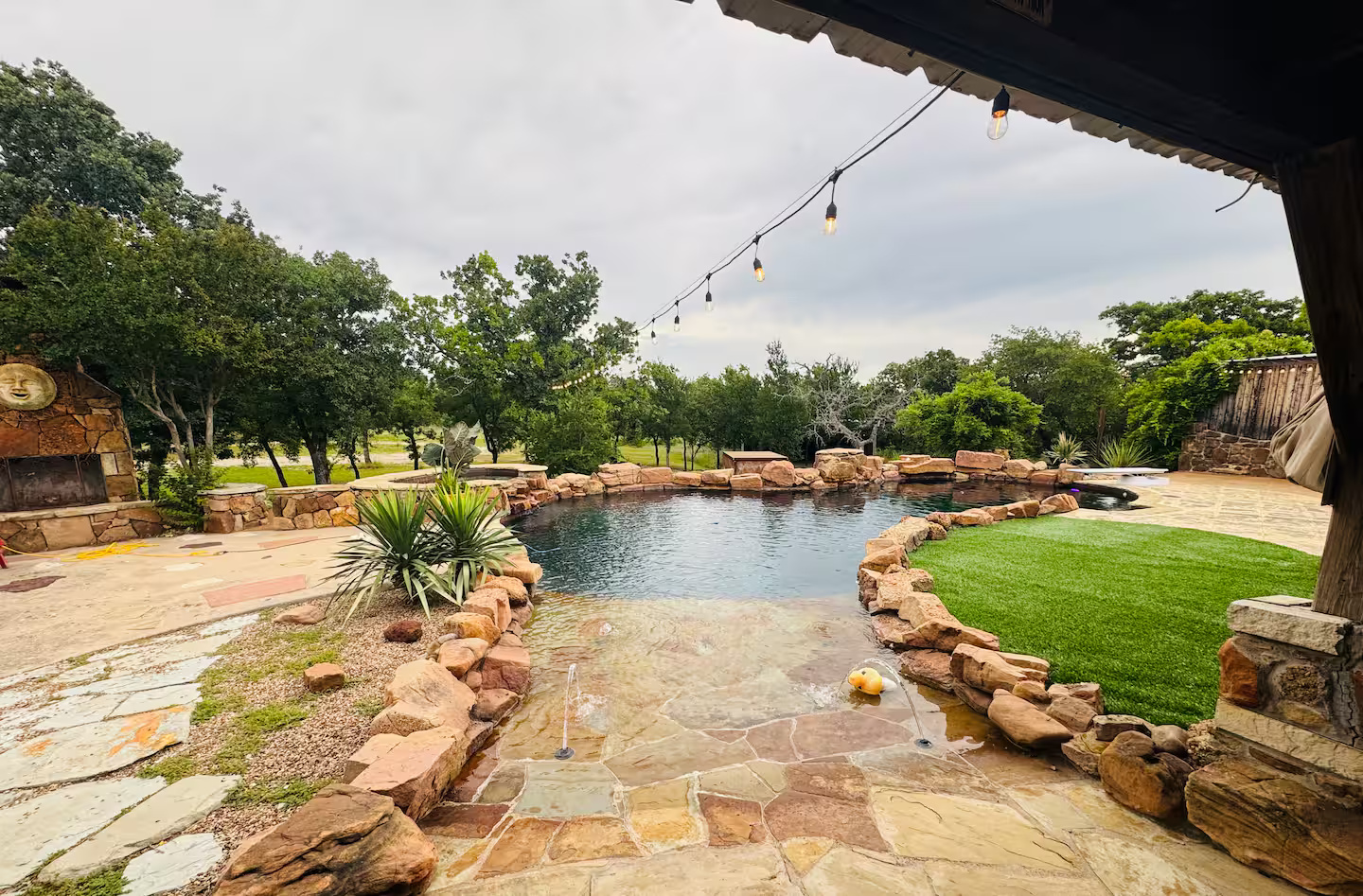 Resort-style pool with shallow stone entry, landscaped stonework, trees, and hanging string lights