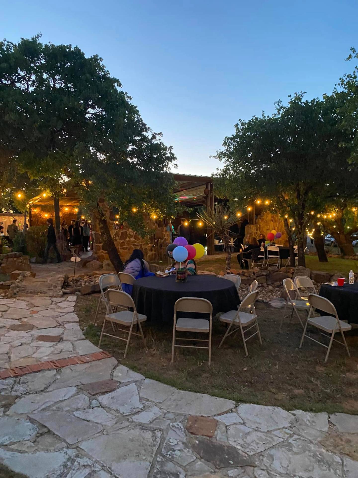 Evening gathering at The Blind Pig — round tables, string lights, stone paths, and balloons under the trees at dusk.