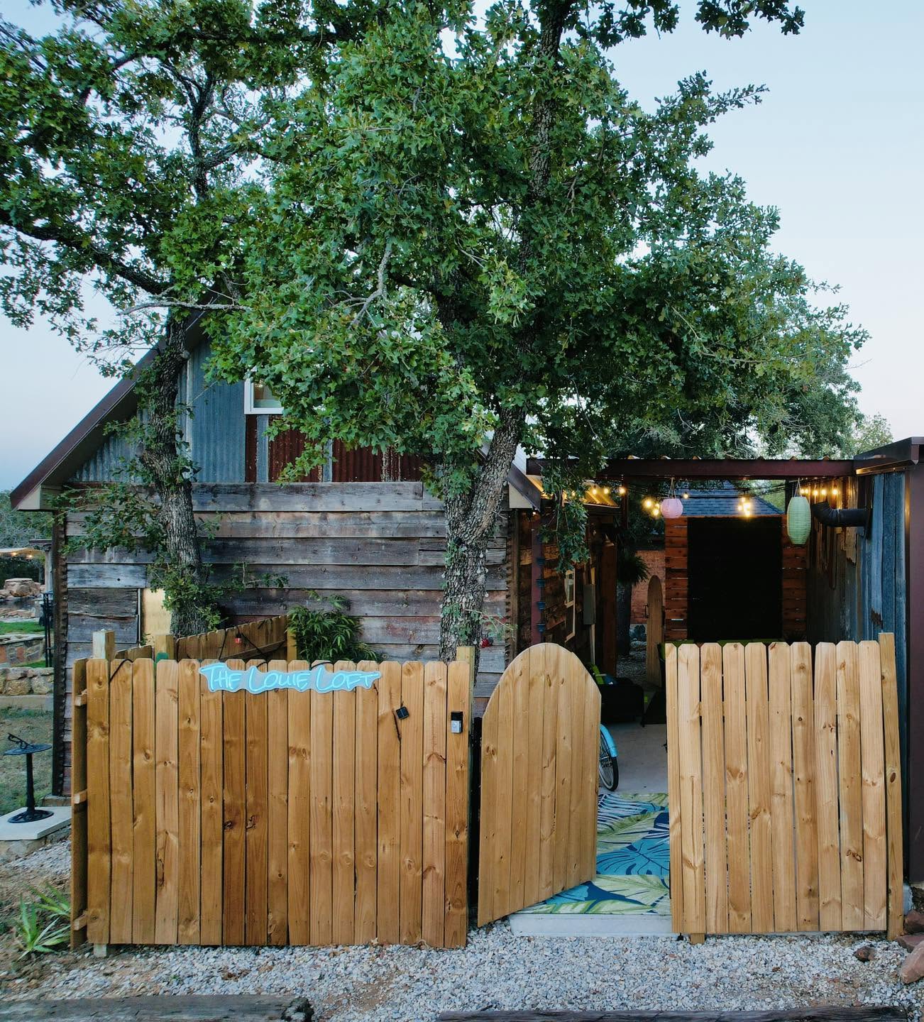 The Louie Loft lodging exterior — reclaimed wood cabin, open wooden gate, oak trees, and warm string lights at dusk.
